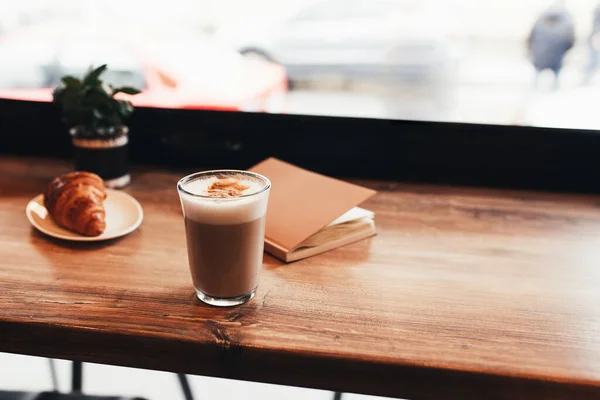 Secangkir kopi, croissant, notebook di meja di kafe. Latar belakang kabur — Stok Foto