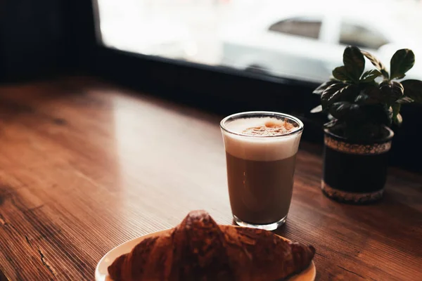 Secangkir kopi, croissant di meja dekat jendela di kafe. Latar belakang kabur — Stok Foto