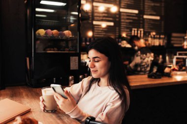 Kız gülümsüyor, kafede kahve içiyor ve telefon okuyor. Bulanık arkaplan