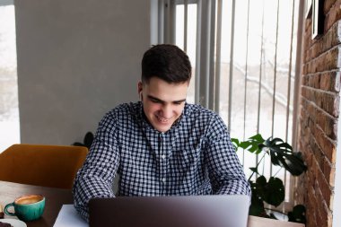 Evde bir fincan kahveyle dizüstü bilgisayarda çalışan gülümseyen adam. Bulanık arkaplan