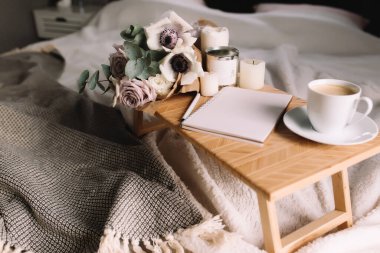 Romantik bir sabah. Yatağında çiçekler olan ahşap sehpa, ekose kahve fincanı, çiçekler ve mumlar, not defteri, kalem. Okaliptüs ve şakayıklı leylak gülleri. İç gri tonlar. Yüksek kalite fotoğraf