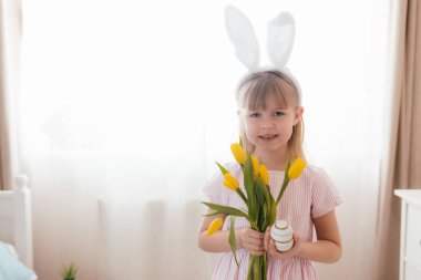 Paskalya konsepti. Beyaz tüylü tavşan kulaklı, elinde sarı lale çiçekleri ve renkli yumurta tutan pembe elbiseli küçük bir kız. İç mekan. Yüksek kalite fotoğraf