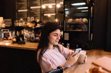 Kız gülümsüyor, kafede kahve içiyor ve telefon okuyor. Arka plan bulanık. Yüksek kalite fotoğraf