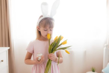 Paskalya konsepti. Beyaz tüylü tavşan kulaklı, elinde sarı lale çiçekleri ve renkli yumurta tutan pembe elbiseli küçük bir kız. İç mekan. Yüksek kalite fotoğraf