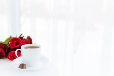 Masada kırmızı lale buketi, beyaz kahve fincanı. Anneler Günü için tebrik kartı kavramı, Uluslararası Kadınlar Günü veya Sevgililer Günü kavramı, doğum günü karşılama kavramı, siyah çikolata şekeri