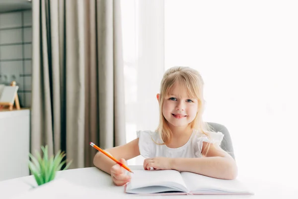 Küçük gülümseyen bir kız masaya oturur ve beyaz bir deftere yazar. Eğitim konsepti. Evde eğitim. Ev ödevi. Gülen surat. Yüksek kalite fotoğraf