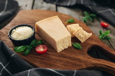 İtalyan mutfağı konsepti. Kesme tahtası, parmesan peyniri parçaları, küçük siyah kasede rendelenmiş peynir, kiraz domatesleri, fesleğen yaprakları, ahşap zeminde mutfak havlusu. Sağlıklı beslenme