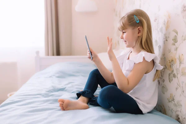 Küçük bir kız şık bir yatak odasında yatağında oturuyor, elinde telefonla bir video görüşmesi yapıyor, akıllı telefon kullanıyor, el sallıyor. İletişim ya da evde eğitim kavramı