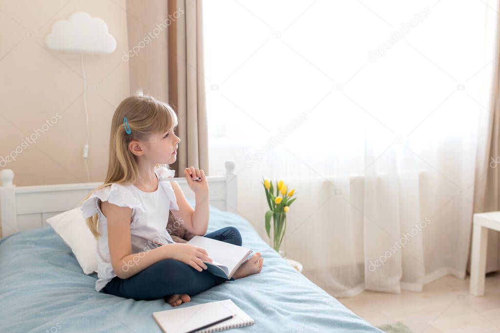 A little girl sits on the bed in the stylish bedroom, holds blue book and pen, doing homework. Thinks about solving a problem. Education and home schooling concept. Thinking on homework