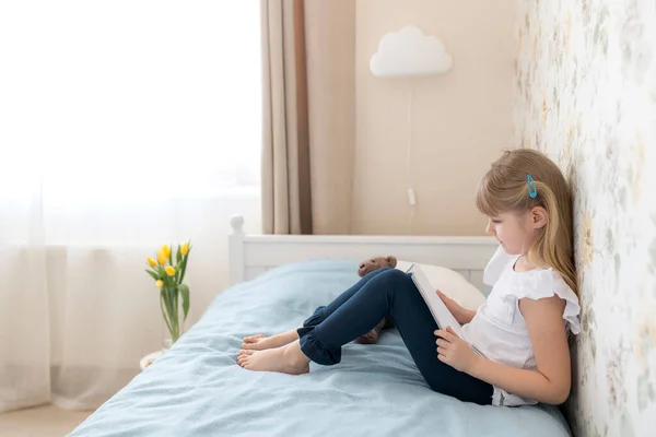 Küçük bir kız şık bir yatak odasında yatağında oturur ve mavi bir kitap okur. Eğitim, evde eğitim kavramı. Ev ödevi. Yatağın yanındaki vazoda sarı laleler var. Bulutlu şamdan.