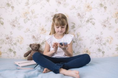 Küçük bir kız şık bir yatak odasında yatağında oturuyor, elinde telefonla bir şeyler okuyor. İletişim konsepti