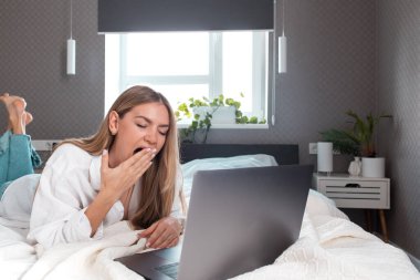 Uzaktan kumandayla yatak odasında dizüstü bilgisayar kullanmaktan bıkmış beyaz bir kız. Uzak çalışma kavramlarının yorgunluğu ve yorgunluğu. Gri iç daire