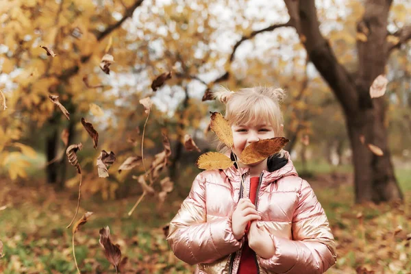 Το ξανθό κοριτσάκι παίζει με κίτρινα φθινοπωρινά φύλλα στον κήπο, χαμογελάει, διασκεδάζει. — Φωτογραφία Αρχείου