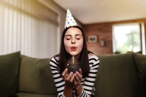 段ボールの帽子の誕生日の女の子のブルネットはソファやソファに座って カップケーキの上で燃えるろうそくを吹き消します 手に誕生日ケーキを持っている女性 背景がぼやけたお祝いのコンセプト — ストック写真