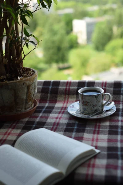 Ekosenin üzerinde porselen bir fincan kahve duruyor, yanında bir kitap yatıyor, ve arka planda pencereden güzel bir manzara var..