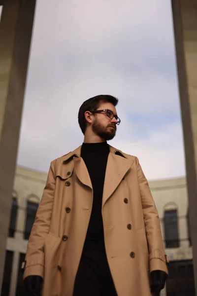 Bej ceketli ve gözlüklü bir adam, kolonlardan oluşan bir kemerle fırtınalı bir gökyüzüne karşı duruyor.