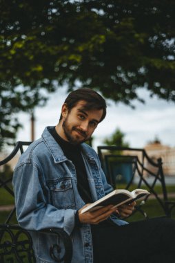 Kot ceketli sakallı bir adam elinde kitapla bankta oturuyor. Arka planda yeşillik ve saray meydanı var..