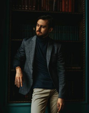 Gri ekose ceketli bir adam kütüphanede duruyor, kitaplığa yaslanıyor.