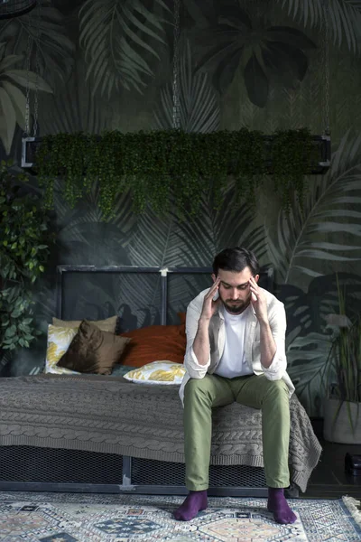 Hafif keten gömlekli bir adam, başı elinde yatakta oturuyor, baş ağrısı var gibi görünüyor.