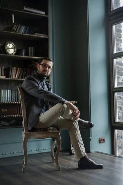 Gri ceketli ve açık yeşil pantolonlu bir adam pencerenin karşısındaki kütüphanede oturuyor.