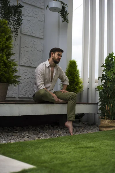 Açık keten gömlekli bir adam yeşil bir terasta oturur.