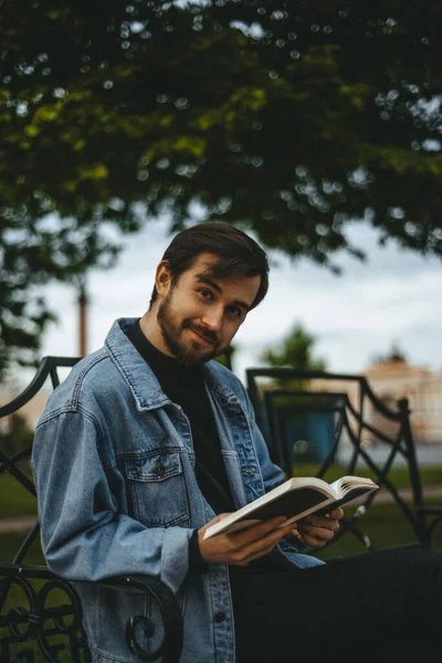 Kot ceketli sakallı bir adam elinde kitapla bankta oturuyor. Arka planda yeşillik ve saray meydanı var..
