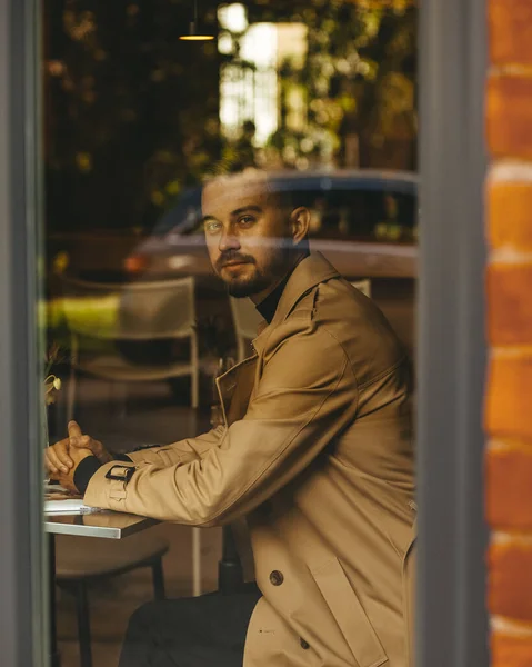 Bej ceketli bir adam kahve dükkanında bir masada oturuyor, ön planda camdan yansıyan bir araba var..