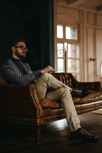 Klasik takım elbiseli bir adam kahverengi deri bir kanepede oturur, arka planda açık bir pencere.