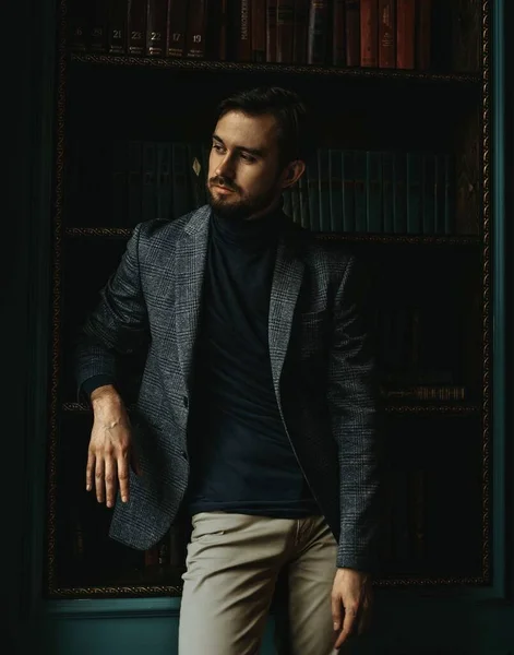 Gri ekose ceketli bir adam kütüphanede duruyor, kitaplığa yaslanıyor.