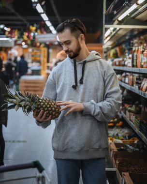 Ön planda, sakallı bir adam elinde bir ananas tutuyor. Arka planda bir süpermarket ve meyveli bir vitrin var..