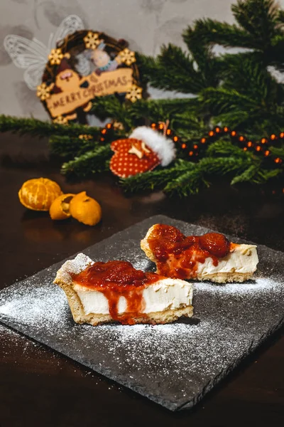 Ön planda çilek reçelli bir cheesecake, arka planda soyulmuş bir mandarin ve kırmızı boncuklu bir ladin dalı ve kırmızı bir eldiven var..