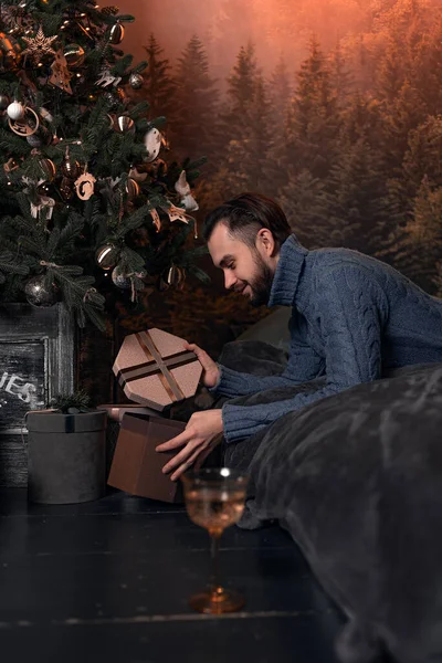 Mavi örgü süveterli bir adam Noel ağacının altında bir hediye kutusu açıyor..