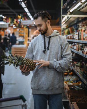 Sakallı bir adam süpermarkette ananas tutuyor.