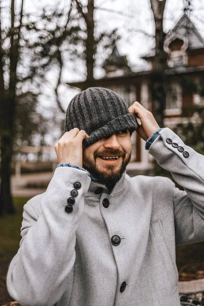 Açık gri ceketli sakallı adam şakacı bir şekilde gri yün bir şapka çekiyor.