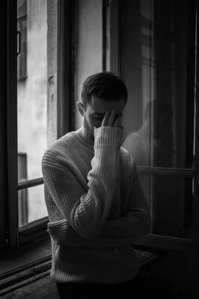 Koyu renk saçlı adam pencerede durur ve elleriyle yüzünü kapatır.