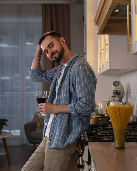 Mavi çizgili gömlekli sakallı adam elinde bir bardak şarapla mutfakta duruyor.