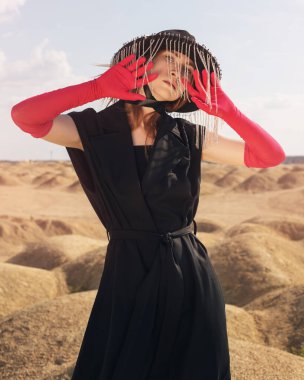 Çölde siyah şapkalı, geniş tarlalı, siyah elbiseli ve kırmızı eldivenli bir kız.