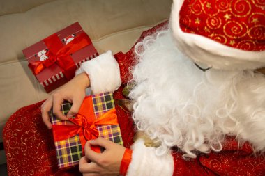 Üst Manzara. Noel Baba gibi giyinmiş bir adam kırmızı kurdeleyle kareli bir kutuya fiyonk bağlıyor. Kanepede oturuyor. Yerde kırmızı bir hediye kutusu var. Yatay. Kapat. Noel zamanı. Yeni yıl. Vintage