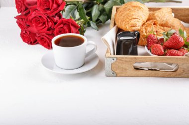 Tahta tepside bir fincan kahve ve kruvasan. Beyaz masanın üzerinde kırmızı güller olan şenlikli bir kahvaltı. Boşluğu kopyala.