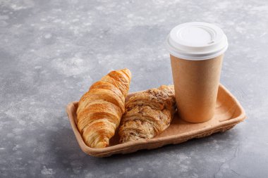 Kahvaltı için sabah kahvesi ve kruvasan. Kahveyi paket yap, kahvaltıyı da götür. Gri masadaki ekmekli kağıt bardak..