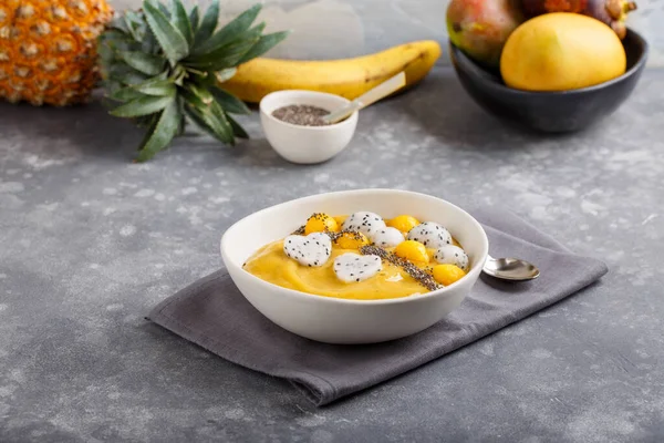 Smoothie bowl on gray background. Delicious mango smoothie topped with chia seeds, pieces of mango and dragon fruit. Healthy breakfast.