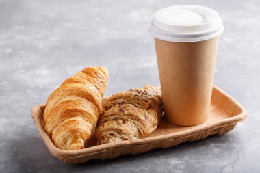 Kahvaltı için sabah kahvesi ve kruvasan. Kahveyi paket yap, kahvaltıyı da götür. Gri masadaki ekmekli kağıt bardak..