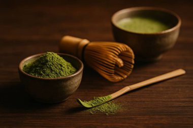 Yeşil çay tozu ile dolu bir kibrit kutusunun yakın çekimi. Hafif pudrayla kaplanmış bambu kepçesinin yanında. Arka planda bir Chasen ve Chawan bulanık görünüyor, derinlik yaratıyor..