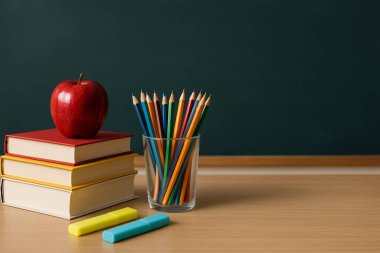 Kırmızı, sarı ve beyaz kaplı kitaplarla dolu bir sınıf masasının yüksek çözünürlüklü fotoğrafı. Üstünde bir elma, renkli kalemler ve floresan kalemler var. Sırtında siyah bir karatahta var.