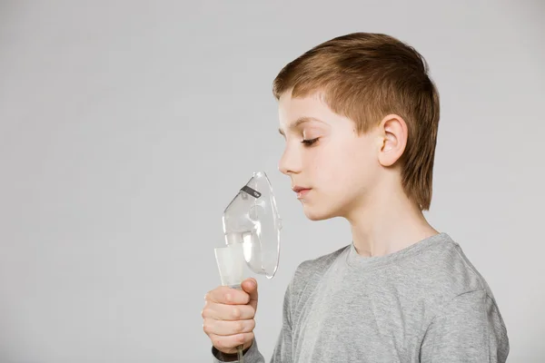 Infelice ragazzo respirando attraverso inalatore maschera su sfondo grigio — Foto Stock