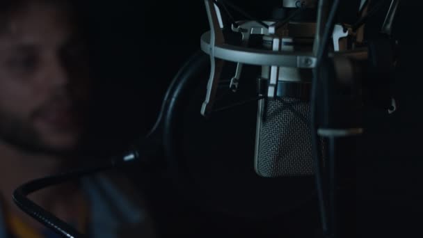 Портрет гарного чоловіка-реп-співака в навушниках з професійним мікрофоном, що співає, записуючи нову пісню в сучасній студії. Зосередьтеся на мікрофоні. Музичне виробництво. Погане світло — стокове відео