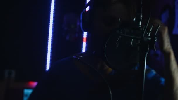 Retrato de un guapo cantante de rap masculino en auriculares con micrófono profesional cantando enérgicamente, grabando nueva canción en un estudio moderno. Producción musical. Mala luz — Vídeos de Stock