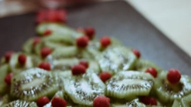 Sulu taze kivi dilimleri ve donmuş kırmızı frenk üzümü siyah mermer kesim tahtasının üzerinde noel ağacı şeklinde düzenlenmiş. Noel tatili için yemek. Sağlıklı atıştırmalıklar. Makro manzara. Yavaş çekim