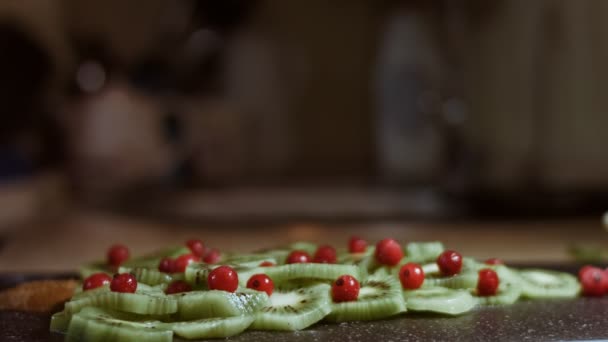 Jugosos trozos de kiwi fresco y grosella roja congelada dispuestos en forma de árbol de Navidad en una tabla de cortar de mármol negro. Verter azúcar en polvo como nieve. Comida para las vacaciones de Navidad. Lento. — Vídeo de stock