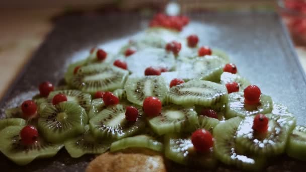 Saftige frische Kiwi-Scheiben und gefrorene rote Johannisbeeren in Form eines Weihnachtsbaums auf einem Schneidebrett aus schwarzem Marmor angeordnet. Pulverzucker gießen, wie es schneit. Essen für die Weihnachtsfeiertage. Zeitlupe — Stockvideo